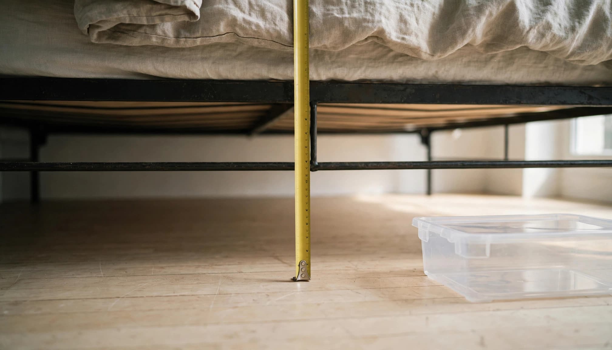 A tape measure checks the space beneath a metal bed beside a clear storage bin.